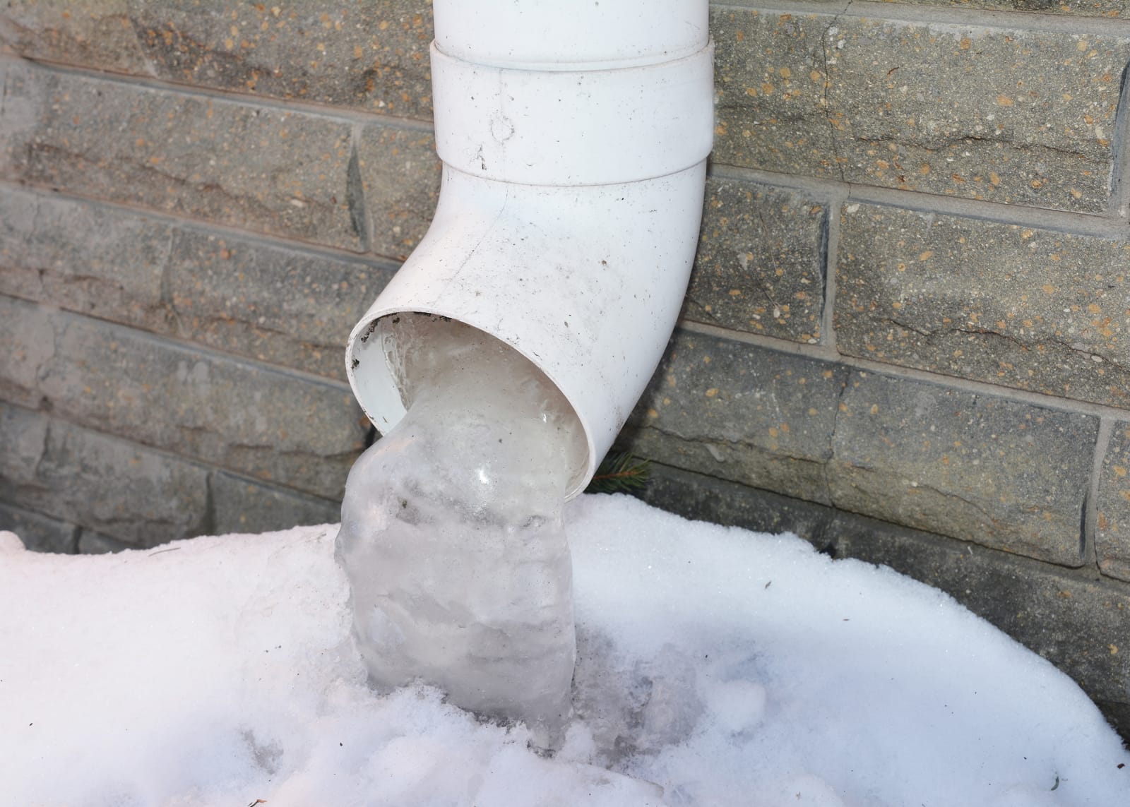 A close-up of a roof gutter downpipe, downspout with frozen water, icicles near the house foundation in winter.