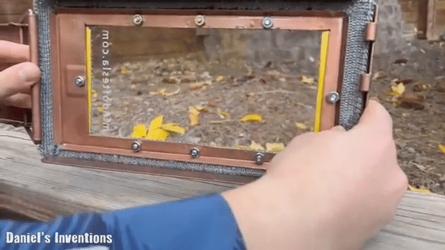 A person is holding a handcrafted, rectangular device with a clear glass front, surrounded by a copper frame with visible screws and mesh edges, labeled with sequences of numbers and letters on the bottom panel. The background visible through the glass is an outdoor scene with autumn leaves.