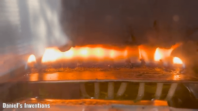 Flames are visible along a metal surface, possibly part of a heating or cooking appliance, under a clear protective cover which has text referring to